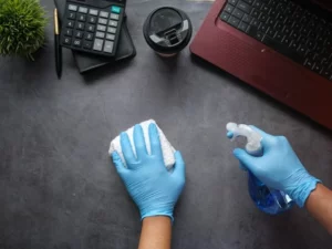 person using cleaning solution to disinfect desk surface wearing blue gloves next to red laptop and coffee cup and calculator